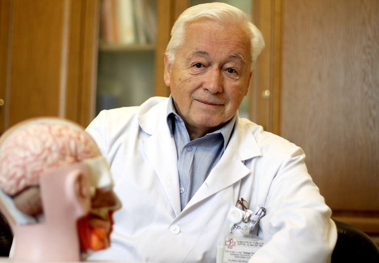 Mężczyzna o siwych włosach i fartuchu lekarskim patrzy w obiektyw aparatu, kt&oacute;ry zrobił zdjęcie. Przed nim model m&oacute;zgu